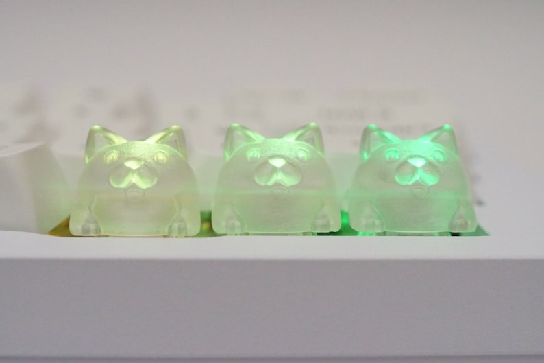 May include: Three translucent green cat-shaped keycaps with a light green glow. The keycaps are arranged in a row on a white surface.