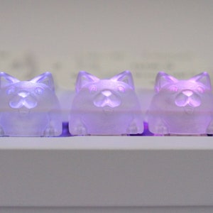May include: Three small, translucent, purple cat-shaped figurines with glowing eyes. The figurines are arranged in a row on a white surface.