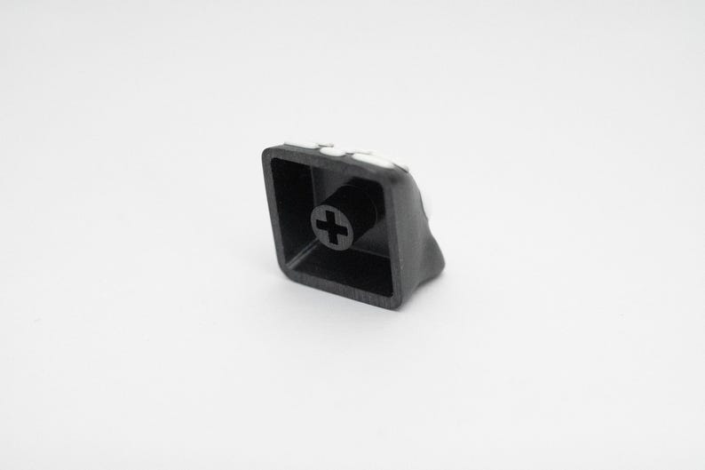 May include: A black plastic keyboard keycap with a plus sign on the top. The keycap has a white border around the edge.