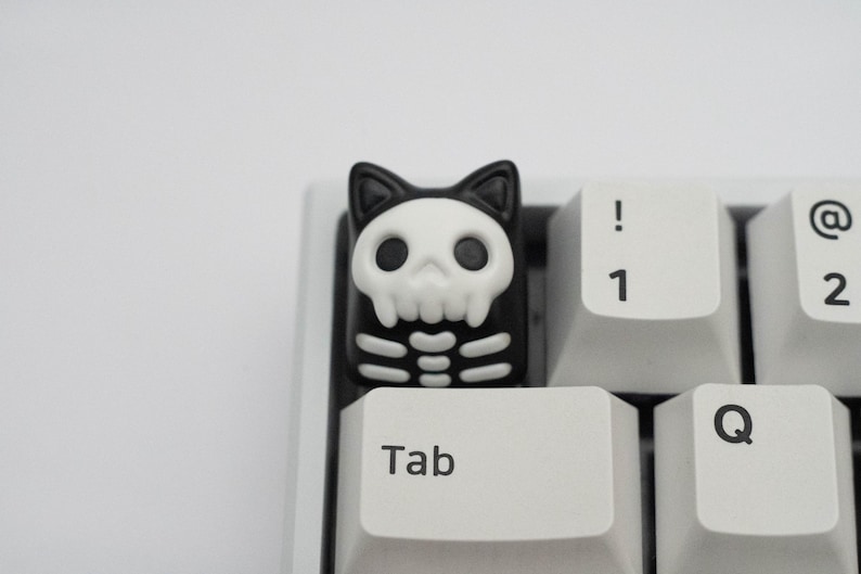 May include: A black and white keyboard keycap with a skull and crossbones design. The keycap is shaped like a cat and has a white skull with black eyes and a black skeleton body. The keycap is on a white keyboard with the keys "Tab", "Q", "1", and "@" visible.