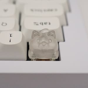 May include: A clear plastic keycap shaped like a cat with a white background. The keycap is sitting on a white keyboard.