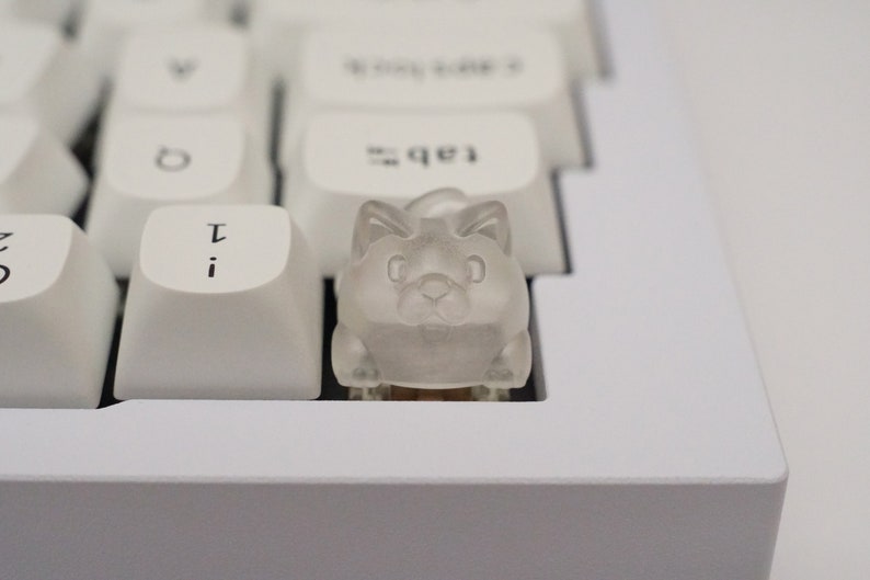 May include: A clear plastic keycap shaped like a cat with a white background. The keycap is sitting on a white keyboard.
