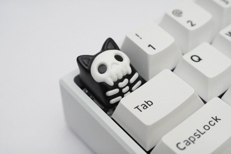 May include: A black and white cat-shaped keycap with a skull and skeleton design. The keycap is on a white keyboard with the keys "Tab" and "CapsLock" visible.