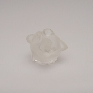 May include: A clear glass figurine of a small animal, possibly a bear or a rabbit, with a rounded body and a small head. The figurine is sitting on a white surface.
