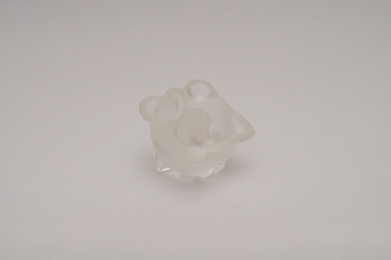 May include: A clear glass figurine of a small animal, possibly a bear or a rabbit, with a rounded body and a small head. The figurine is sitting on a white surface.