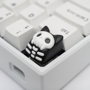 May include: A black and white skeleton cat keycap with a skull design. The keycap is on a white keyboard.