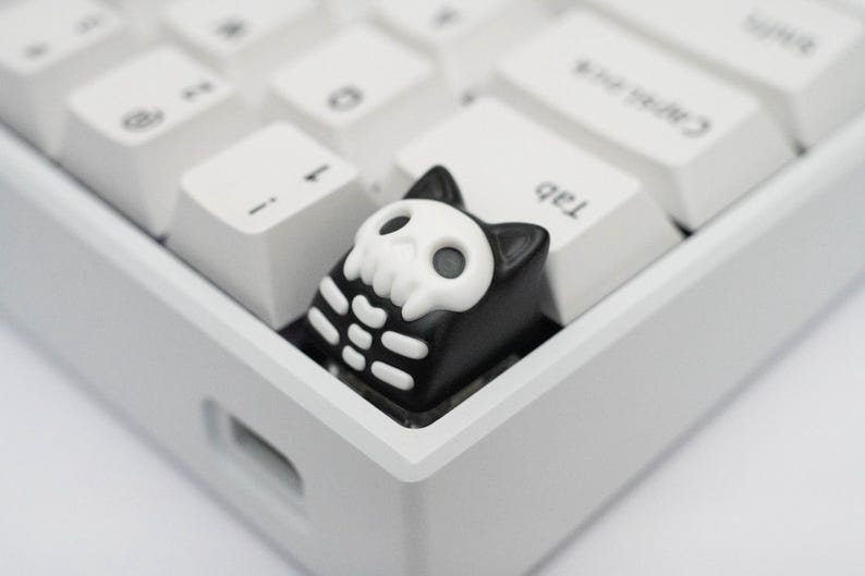 May include: A black and white skeleton cat keycap with a skull design. The keycap is on a white keyboard.
