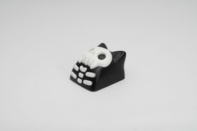 May include: Black and white keycap with a skull design and a cat-like shape.