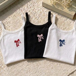 May include: Three vest tops: two white and one black. Each top features bow and initial embroidery. The white tops have red and blue bows, while the black top has a pink bow. The tops are on a textured surface.
