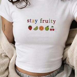 May include: A white short-sleeved t-shirt featuring the embroidered text stay fruity in multicoloured letters. Below the text are small embroidered icons of a red strawberry, a green lime wedge, an orange slice, a green apple, and two red cherries.