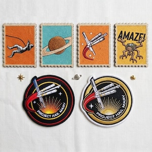 Könnte beinhalten: Gestickte Aufnäher und Briefmarken mit Weltraum-Designs. Die Briefmarken zeigen einen Astronauten, Saturn, ein Raumschiff und einen Außerirdischen mit dem Wort "AMAZE!". Zwei runde Aufnäher tragen die Aufschrift "PROJECT HAIL MARY".