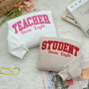 May include: Two sweatshirts, one white and one beige, are displayed. The white sweatshirt has "TEACHER" embroidered in pink with "Laura Kayla" below. The beige sweatshirt has "STUDENT" in red with "Laura Kayla" below. Both have a crew neck.