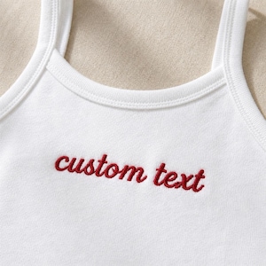 May include: White vest top with red embroidered text reading "custom text". The top features a scoop neckline and thin straps. The fabric appears to be a soft, knit material.