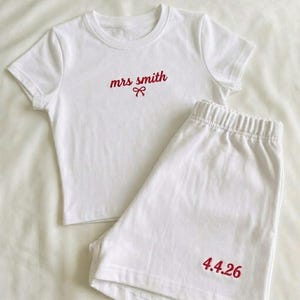 Op de afbeelding: Witte pyjamaset met een top met korte mouwen en bijpassende shorts. De top is geborduurd met "mrs smith" in rode letters met een strik. De shorts zijn geborduurd met de datum "4.4.26" in rood.
