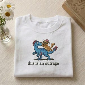 May include: White t-shirt featuring an embroidered design of a blue dinosaur and a cat wearing a crown. The cat is riding the dinosaur. The text "this is an outrage" is below the graphic.
