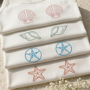 Puede incluir: Una pila de bodies blancos para bebés con diseños bordados con temática marina. Los diseños incluyen conchas rosas, caracolas verdes, dólares de arena azules y estrellas de mar coral. Los bodies están cuidadosamente doblados y apilados.