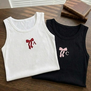 Op de afbeelding: Twee mouwloze tanktops, één witte en één zwarte, worden getoond. De witte tanktop heeft een rode strik en de letter "A" geborduurd. De zwarte tanktop heeft een roze strik met de letter "C". Beide tops zijn gemaakt van zacht materiaal.