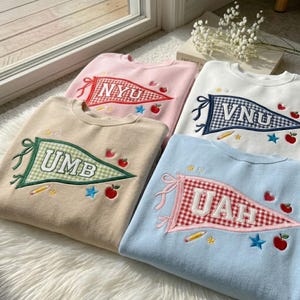 Op de afbeelding: Vier crewneck sweatshirts in pastelkleuren: roze, beige, wit en lichtblauw. Elk heeft een geborduurd wimpelontwerp met letters, sterren, appels en potloden. De sweatshirts liggen op een witte, zachte ondergrond.