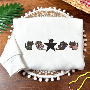 Peut inclure: Sweat-shirt blanc avec un motif de cinq chats de dessin animé. Les chats sont dans différentes poses, dont un sur une étoile noire. Le sweat-shirt a un col rond et des manches longues. Le motif utilise du marron, du vert et du rose.