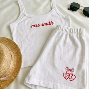 May include: White pajama set comprising a vest top and shorts. The vest top is embroidered with "mrs smith" in red. The shorts feature two red embroidered hearts with the letters "P" and "L" and a bow. A straw hat and sunglasses are also in the image.