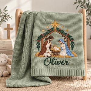 May include: A sage green knitted blanket featuring an embroidered nativity scene with a central star, a manger, and the name Oliver stitched in dark green script. The design includes figures in brown and blue robes, a lamb, and greenery.