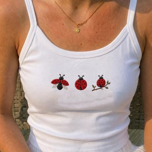 May include: A white vest top featuring three embroidered ladybird designs across the chest. The first ladybird is shown with wings spread, the second is circular, and the third is perched on a small green branch. The top has a scoop neckline.