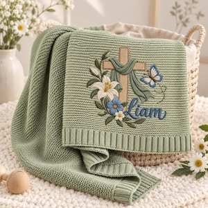 May include: A sage green knitted blanket featuring an embroidered design of a tan cross draped with green fabric, white lilies, blue flowers, a blue butterfly, and the name Liam stitched in blue script, finished with a ribbed border edge.