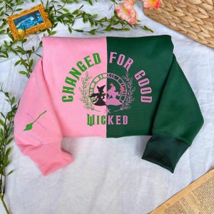 Puede incluir: Una sudadera de dos tonos, dividida verticalmente con rosa y verde oscuro. El lado rosa presenta un gráfico de escoba verde. El lado verde tiene un diseño circular con dos siluetas de brujas y las palabras "CHANGED FOR GOOD" y "WICKED".