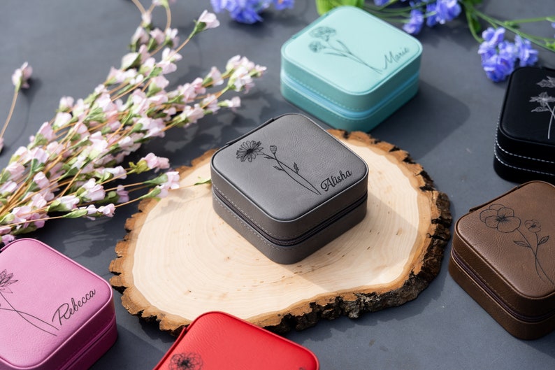 May include: Six small, square, personalised jewellery boxes with floral designs. The boxes are in various colours, including pink, blue, grey, brown, and black. Each box has a different name engraved on it: Rebecca, Maria, Alisha, and three others.