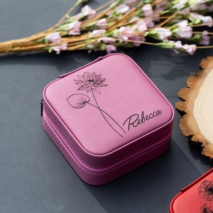 May include: A pink square-shaped jewellery box with a zipper closure. The box has a floral design and the name "Rebecca" engraved on the front.