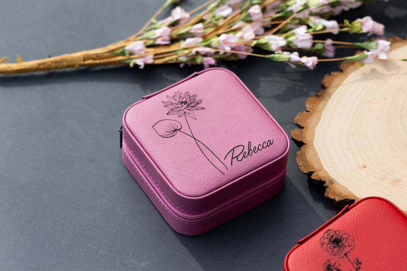 May include: A pink square-shaped jewellery box with a zipper closure. The box has a floral design and the name "Rebecca" engraved on the front.