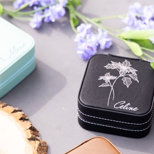 May include: Three small black, brown, and light blue jewellery boxes with floral designs and personalised names. The boxes are on a grey surface with a wooden round slice and purple flowers.