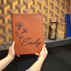 May include: A rectangular tan leather portfolio featuring a black line art illustration of two flowers and the name Emily printed in a cursive script. The cover has a smooth, textured surface and a stitched border along the edges of the front panel.
