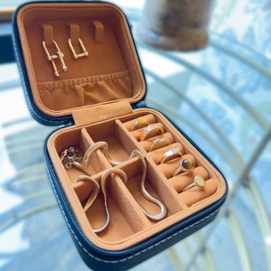 May include: A black leather jewellery box with a brown velvet interior. The box has multiple compartments for storing rings, earrings, and a necklace. The box is open and the compartments are filled with gold jewellery.