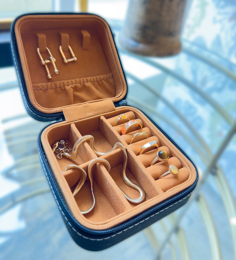 May include: A black leather jewellery box with a brown velvet interior. The box has multiple compartments for storing rings, earrings, and a necklace. The box is open and the compartments are filled with gold jewellery.