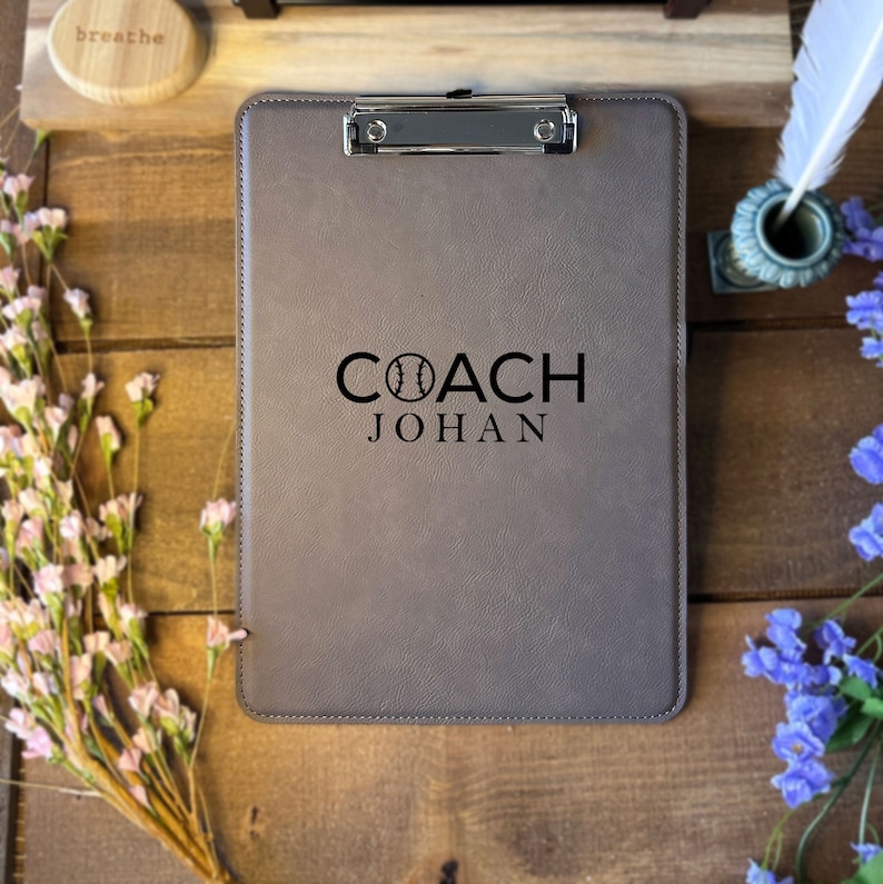 Może przedstawiać: Szary klips z srebrnym klipsem, z czarnym napisem "COACH JOHAN". Słowo "COACH" zawiera grafikę baseballową. Klips leży na drewnianej powierzchni, z drewnianym dyskiem z napisem "breathe".