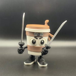 Pode incluir: Um personagem de xícara de café impresso em 3D com tema ninja. A xícara tem uma tampa marrom, uma faixa bege e um corpo branco. Ele está usando uma bandana e segurando duas katanas prateadas. O personagem tem sapatos e braços pretos.