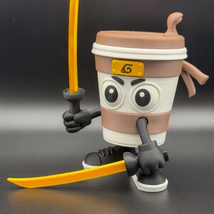 May include: A 3D-printed coffee cup figure with a cartoonish design, holding two yellow swords. The cup has a brown lid, a white body with a brown band, and black gloves and shoes. A yellow emblem is on the cup.