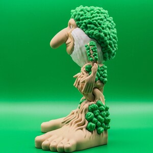 May include: A quirky figurine depicting a bearded character with a prominent nose and green, textured hair. The figure has a light brown complexion and is decorated with green foliage. The base of the figure is shaped like large feet.