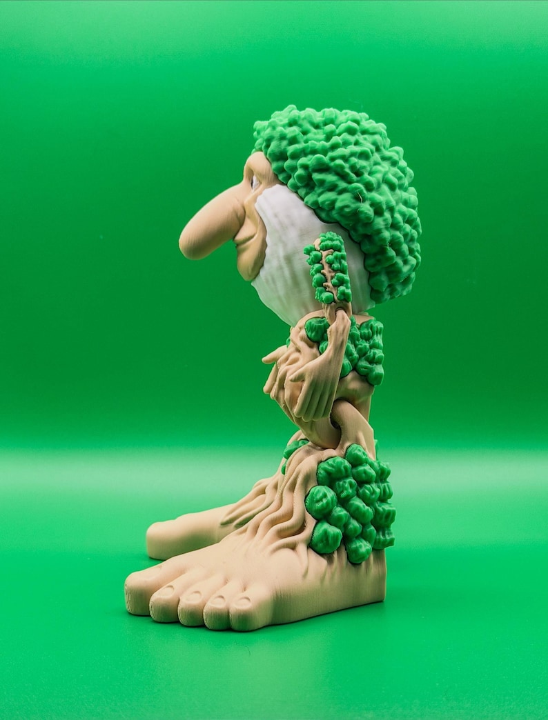 May include: A quirky figurine depicting a bearded character with a prominent nose and green, textured hair. The figure has a light brown complexion and is decorated with green foliage. The base of the figure is shaped like large feet.