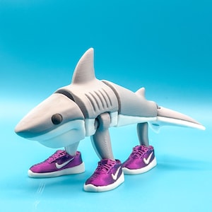 May include: A 3D-printed shark figurine in shades of gray and white, sporting purple sneakers with white swooshes. The shark has a fin, gills, and a tail, and is posed on a light blue surface.