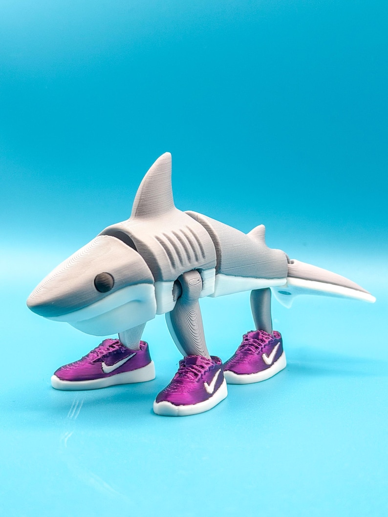 May include: A 3D-printed shark figurine in shades of gray and white, sporting purple sneakers with white swooshes. The shark has a fin, gills, and a tail, and is posed on a light blue surface.