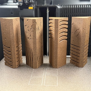 Peut inclure: Quatre piliers triangulaires de couleur brune avec une texture granuleuse. Chaque pilier présente des motifs sculptés distincts sur ses faces avant, notamment des lignes horizontales, des vagues verticales et des textures de surface fissurées.