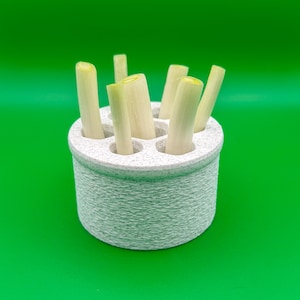 May include: A white speckled ceramic holder with multiple holes, filled with fresh, light green leeks. The holder has a textured, woven appearance. The leeks are cut to various lengths, showcasing their fresh, green and white colors.