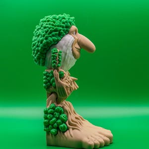 May include: A quirky figurine of a troll-like character with a prominent nose, green hair, and a beard. The figure has a tree-like body with green foliage details and a large foot. The background is a solid green.