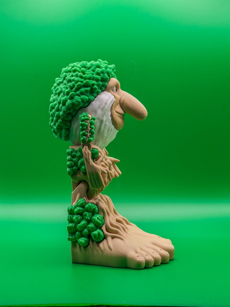 May include: A quirky figurine of a troll-like character with a prominent nose, green hair, and a beard. The figure has a tree-like body with green foliage details and a large foot. The background is a solid green.