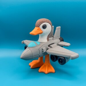 May include: A whimsical toy duck with a jet plane body. The duck has a brown head, white feathers, and an orange beak and feet. The gray jet plane has a blue cockpit and black wheels, set against a blue background.