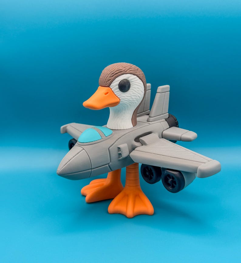 May include: A whimsical toy duck with a jet plane body. The duck has a brown head, white feathers, and an orange beak and feet. The gray jet plane has a blue cockpit and black wheels, set against a blue background.
