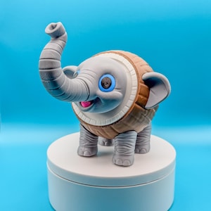 May include: A whimsical, gray elephant figurine with a trunk raised high. The elephant has large blue eyes, a pink tongue, and a brown and tan patterned body. It stands on a white, round platform against a blue backdrop.