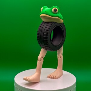 May include: A whimsical sculpture of a frog head atop a black tire, with articulated legs and feet. The frog is green with yellow eyes and a tan underside. The sculpture is on a white platform against a green backdrop.